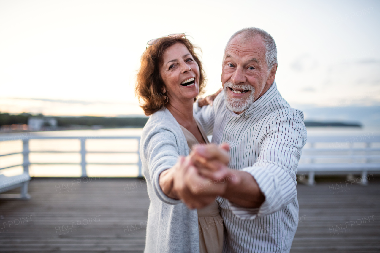 A happy senior couple dancing outdoors on pier by sea, looking at camera.