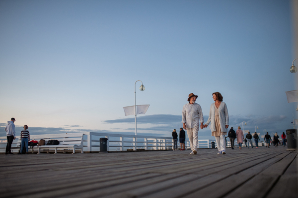 Low angle view of a happy senior couple in love on walk outdoors on pier by sea at dusk, holding hands.