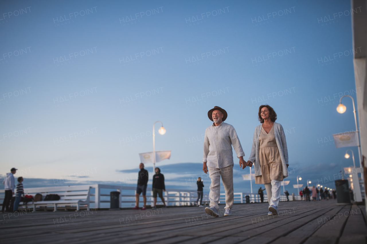 A low angle view of happy senior couple walking outdoors on pier by sea at dusk, holding hands.