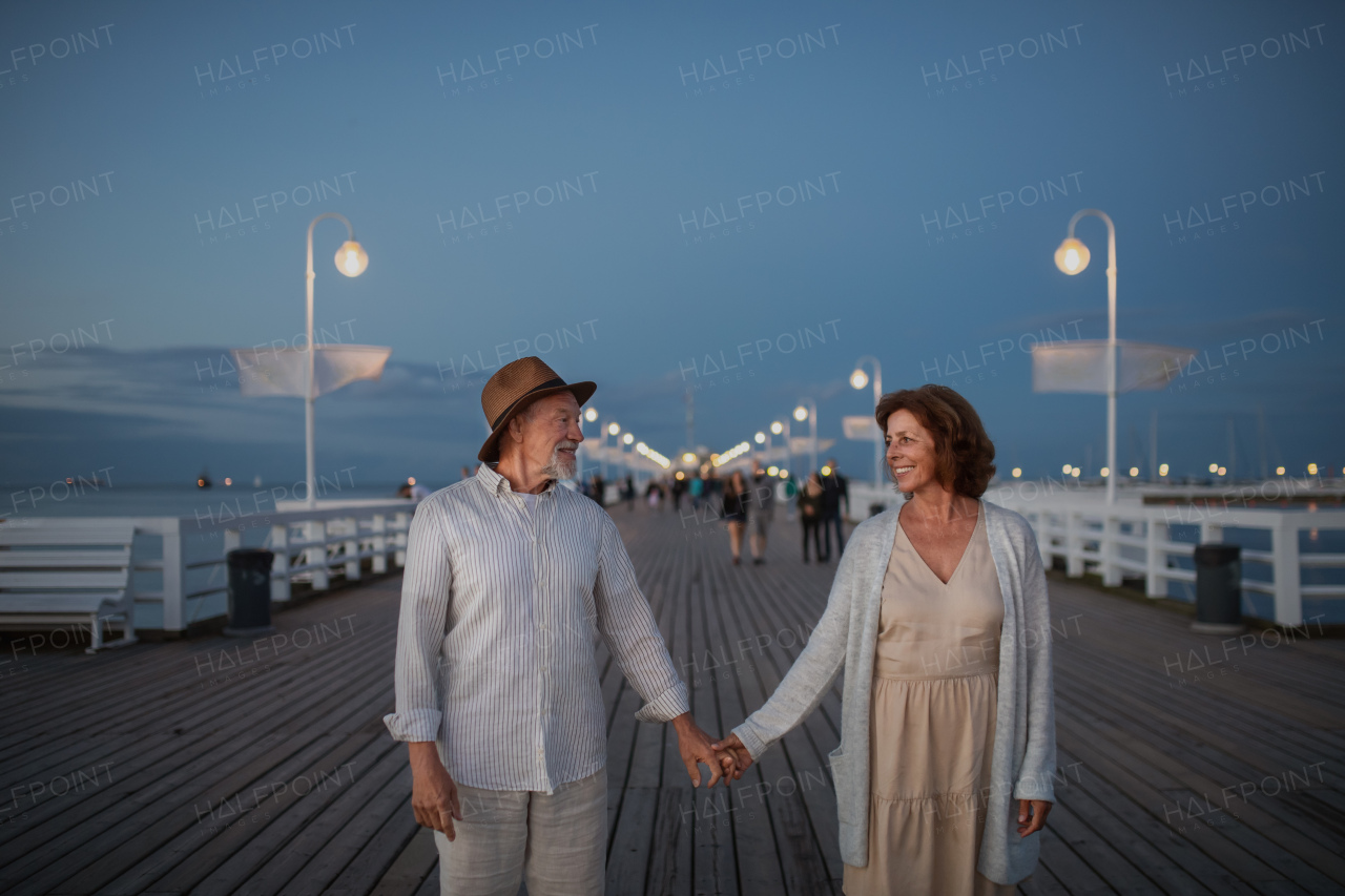 A front view of happy senior couple walking outdoors on pier by sea at dusk, holding hands.