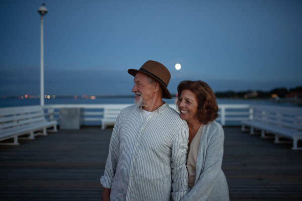 A happy senior couple in love on walk holding hands outdoors on pier by sea at moonlight, looking at view.