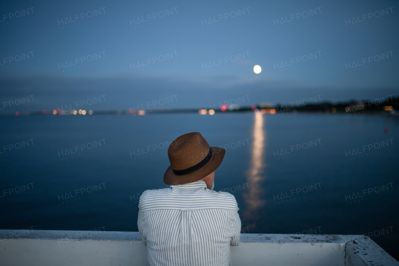 Rear view of senior man tourist loooking at moon outdoors on pier by sea in the evening.