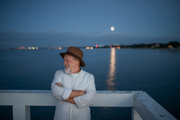 A portrait of senior man tourist loooking aside outdoors on pier by sea.