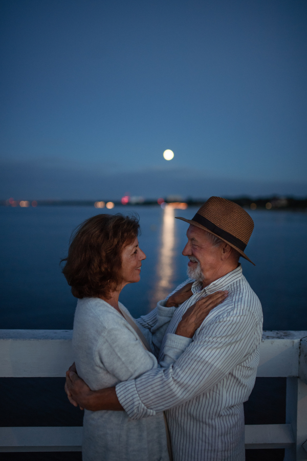 A happy senior couple in love hugging outdoors on pier by sea at sunset, looking at each other.