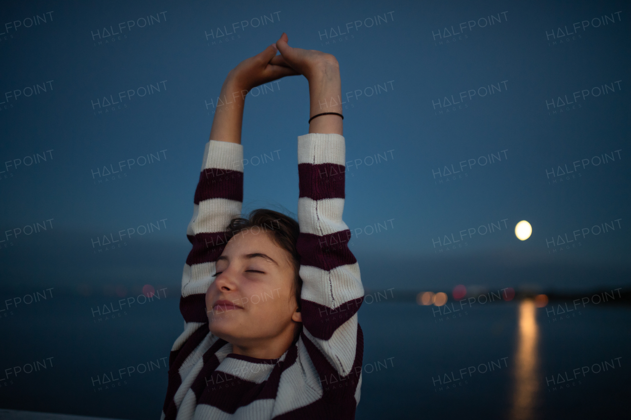 Portrait of a preteen girl stretching with eyes closed outdoors on pier by sea at dusk, holiday concept.