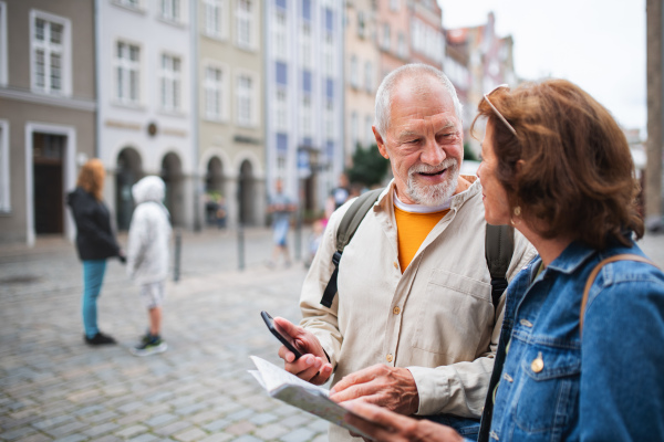 A portrait of happy senior couple tourists using map and smartphone outdoors in town street