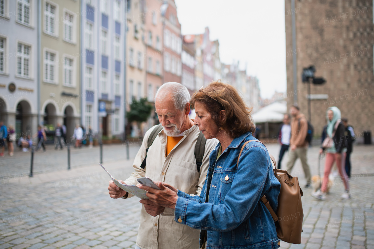 A portrait of senior couple tourists using map and smartphone outdoors in town street