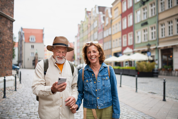 A portrait of happy senior couple tourists smiling, holding hands, using smartphone outdoors in historic town