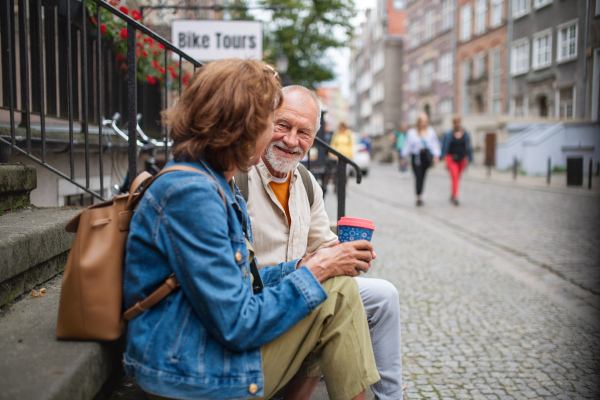 A happy senior couple tourists sitting on stairs and having take away coffee outdoors in town