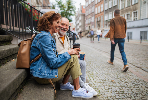 A happy senior couple tourists sitting on stairs and having take away coffee outdoors in town