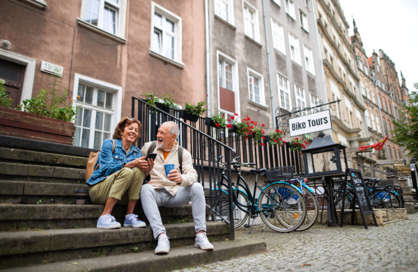 A happy senior couple tourists sitting on stairs and having take away coffee outdoors in town