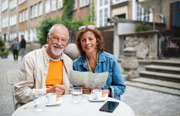 A portrait of happy senior couple tourists sitting and using map outdoors in town, looking at camera.