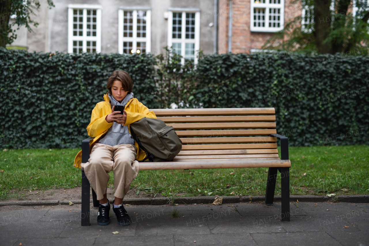 A preteen schoolgirl sitting on bench and using smartphone outdoors in town.