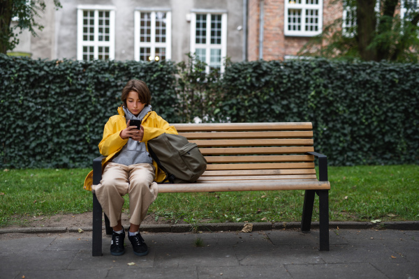 A preteen schoolgirl sitting on bench and using smartphone outdoors in town.