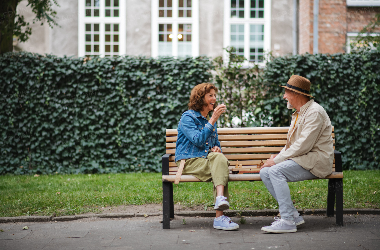 A happy senior couple sitting on bench and playing chess outdoors in park.