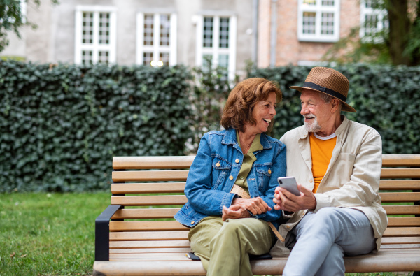 A happy senior couple in love sitting on bench and using smartphone outdoors in town, copy space.