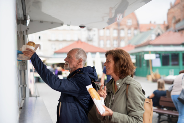 A happy senior couple tourists buying snack outdoors in street.