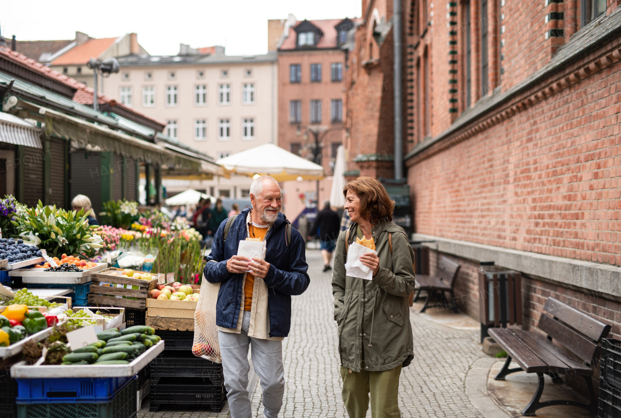 A happy senior couple tourists with snack in town on outdoor market.