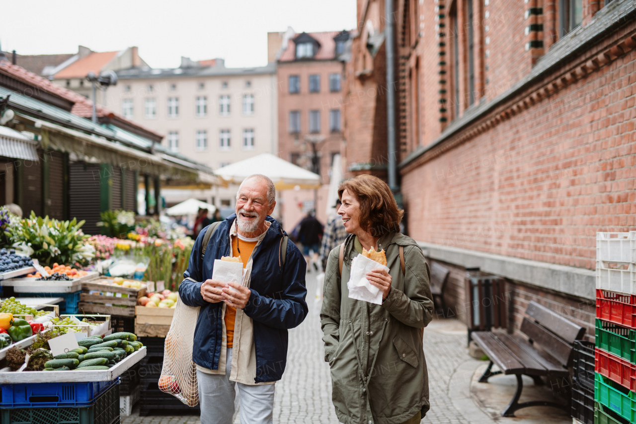 A happy senior couple tourists with snack in town on outdoor market.