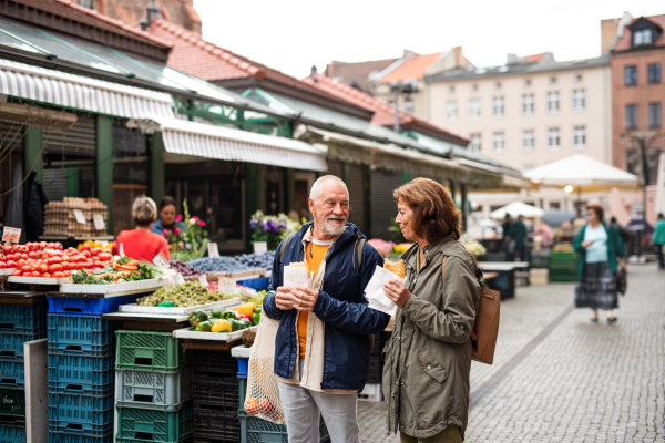 A portrait of happy senior couple tourists with snack in town on outdoor market.