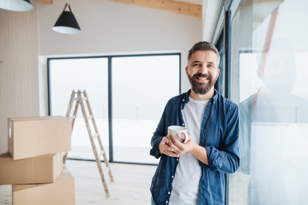 A mature man with cardboard boxes and cup of coffee standing by a window, furnishing new house. A new home concept. Copy space.