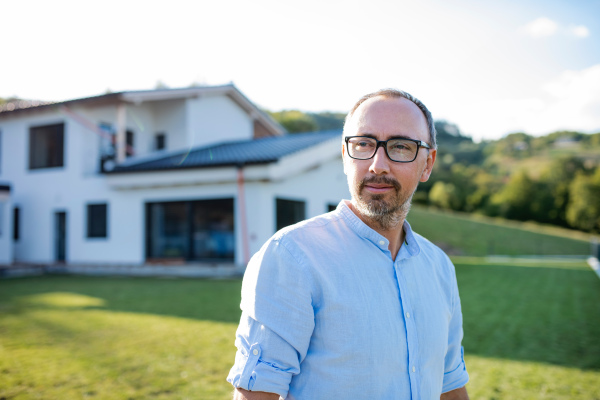 A front view portrait of man standing outdoors in garden by a house.