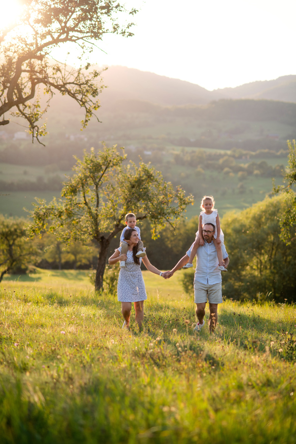 Front view of family with two small children walking on meadow outdoors at sunset, piggyback ride.