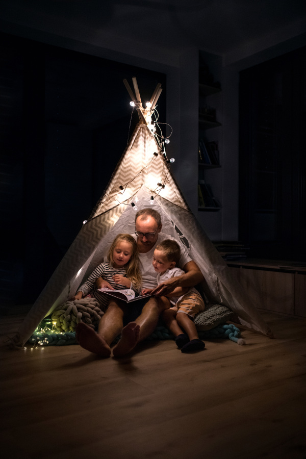 Father with small children sitting indoors in tent in bedroom in the evening, reading a book.