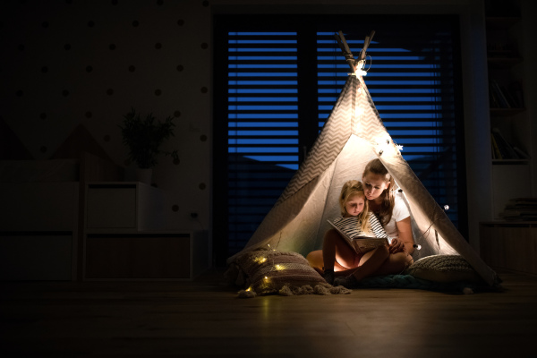 Young mother with small daughter sitting indoors in bedroom, reading a book.