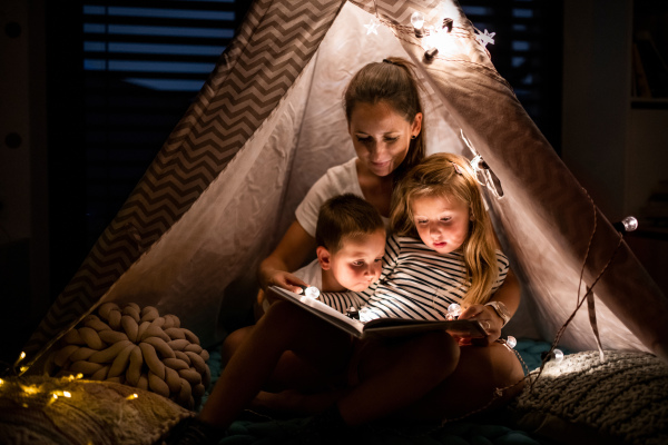 Mother with small children sitting indoors in tent in bedroom in the evening, reading a book.