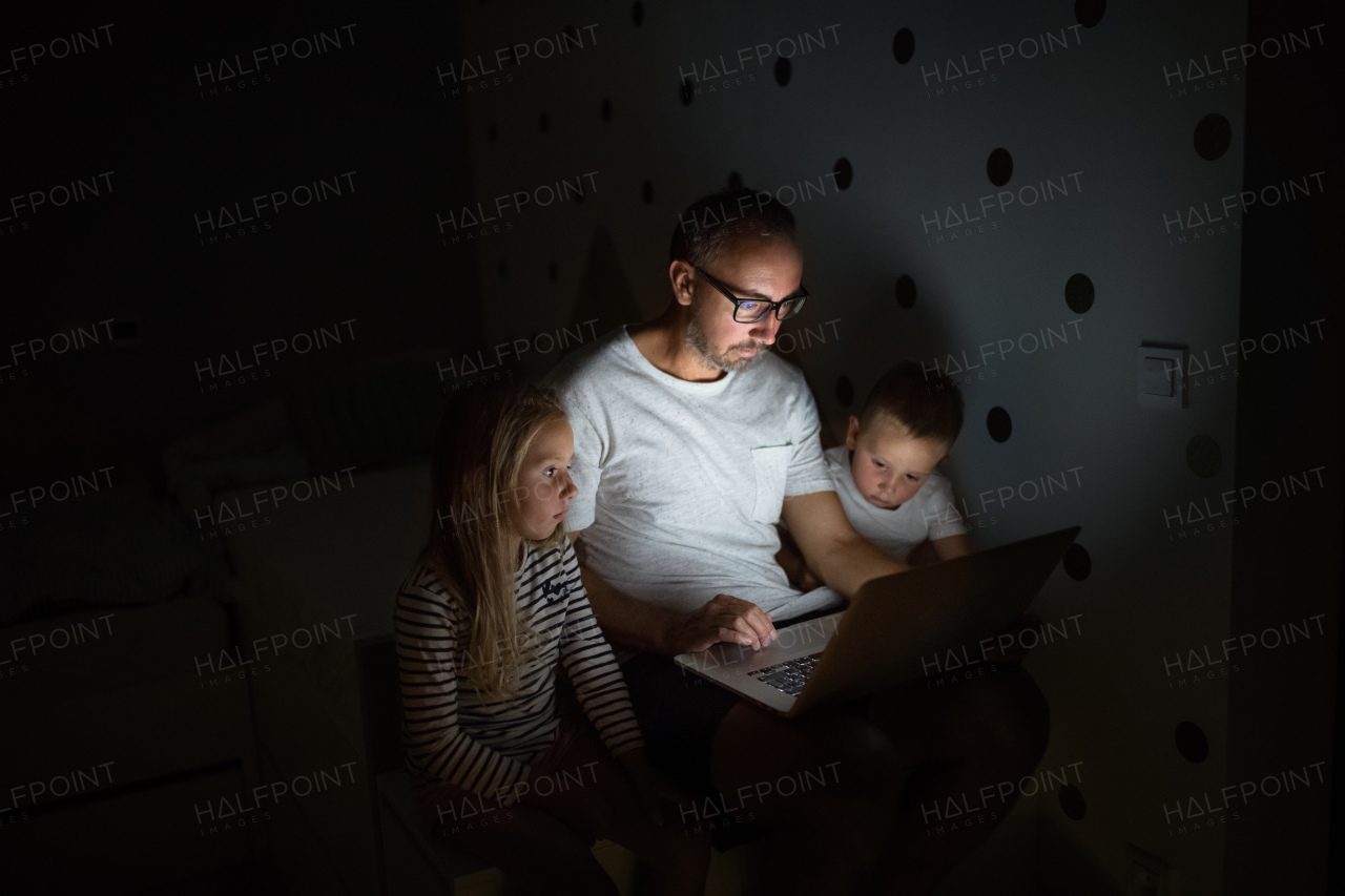 Father with small children sitting indoors in bedroom in the evening, using laptop.