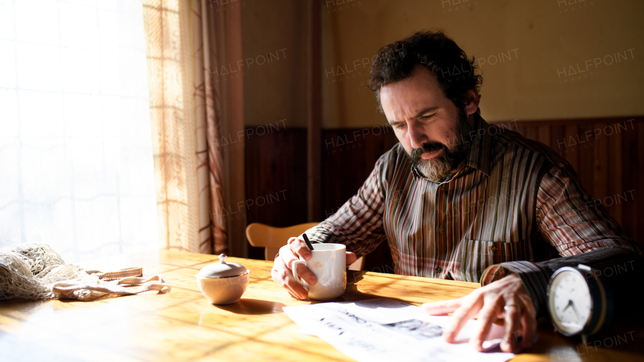 Portrait of unhappy poor mature man with coffee reading newspapers indoors at home, poverty concept.