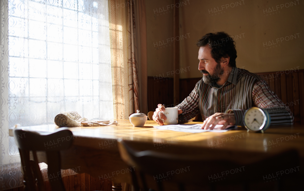 Portrait of unhappy poor mature man with coffee reading newspapers indoors at home, poverty concept.