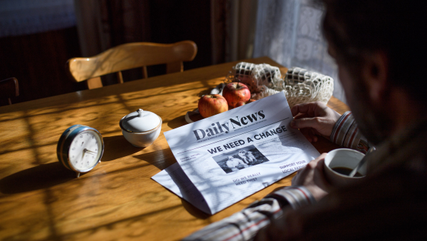 An unrecognizalbe poor mature man reading newspapers indoors at home, poverty concept.