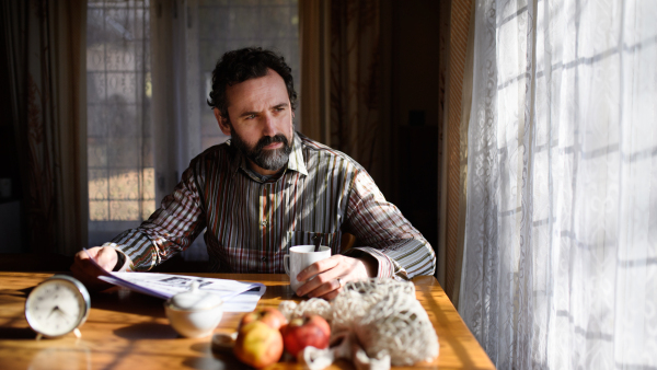 Portrait of unhappy poor mature man with coffee reading newspapers indoors at home, poverty concept.