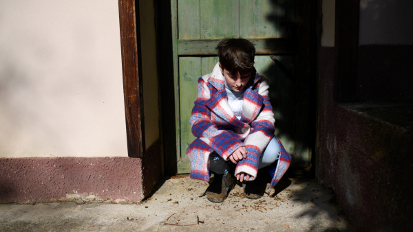 A poor sad small girl with large coat outdoors in front of house, poverty concept.