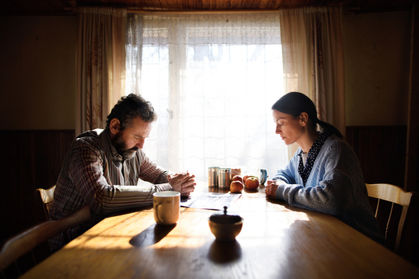 A portrait of sad poor mature couple praying at the table indoors at home, poverty concept.