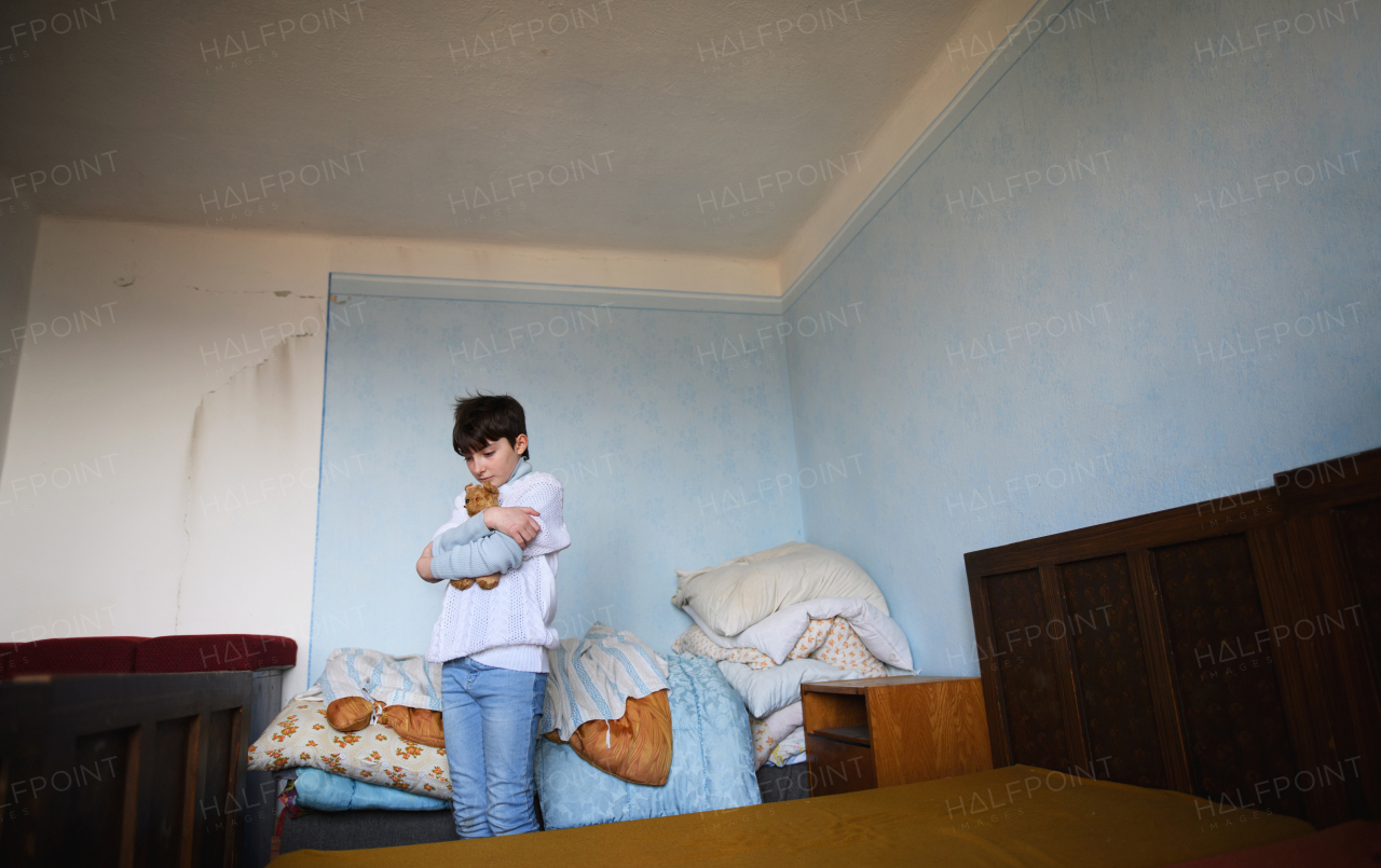 Poor sad small girl sitting in bedroom indoors at home, poverty concept.