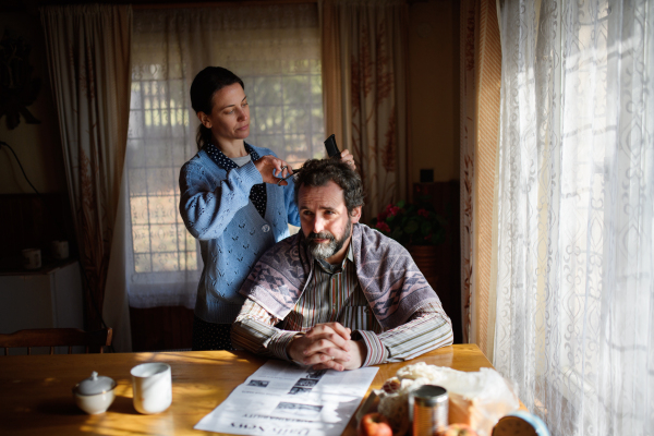 A portrait of poor woman cutting husbands hair indoors at home, poverty concept.