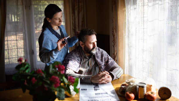 A portrait of poor woman cutting husband's hair indoors at home, poverty concept.