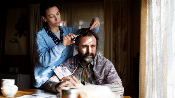 A portrait of poor woman cutting husbands hair indoors at home, poverty concept.