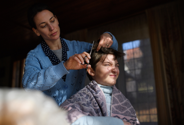 Portrait of sad poor woman cutting daughter's hair indoors at home, poverty concept.