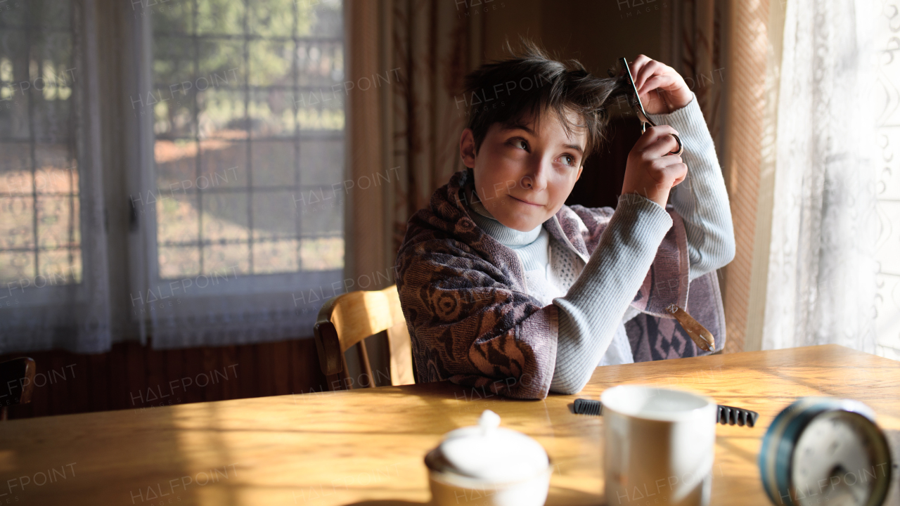 Poor sad small girl sitting at the table and cutting hair indoors at home, poverty concept.