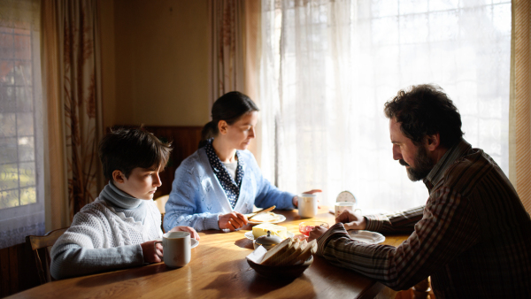 A portrait of poor sad small girl with parents eating indoors at home, poverty concept.