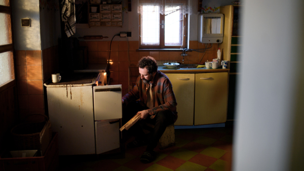 Poor mature man putting firewood in the wood burning stove indoors at home, poverty concept.