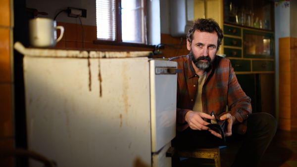 Portrait of poor mature man with empty wallet looking at camera indoors at home, poverty concept.