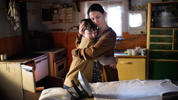 A portrait of sad poor mature mother hugging small daughter indoors at home, poverty concept.