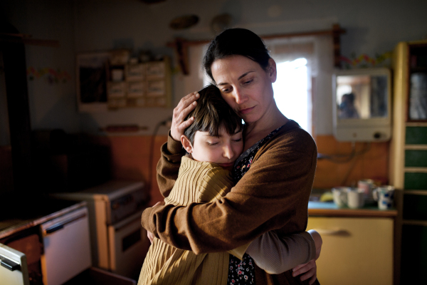 A portrait of sad poor mature mother hugging small daughter indoors at home, poverty concept.