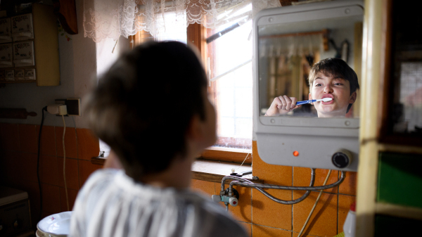 Small girl brushing teeth and looking in mirror in kitchen indoors at home, poverty concept.