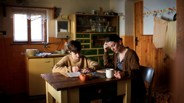 A poor mature mother and small daughter learning indoors at home, poverty concept.